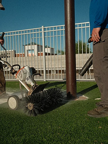 Close view of a power broom head lifting sand out of artificial turf around a light pole base